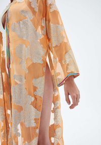 Průhledný oranžový háv se stříbrnou květinovou výšivkou a mnohobarevným lemem, nošený rozepnutý, aby odhalil jednu nohu a bílý oděv pod ním.