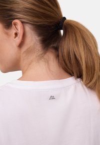 Witte katoenen T-shirt met een ronde hals. Heeft een klein grijs driehoekig logo op de achterkant. Glad van textuur, eenvoudig ontwerp.