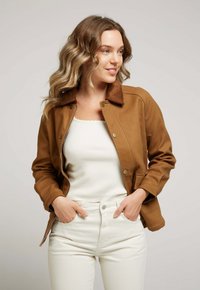 Veste brune avec un col en velours côtelé, fermeture à boutons et deux poches latérales, portée sur un haut blanc et un jean clair.