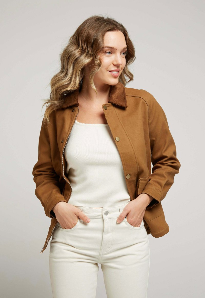 Veste brune avec un col en velours côtelé, fermeture à boutons et deux poches latérales, portée sur un haut blanc et un jean clair.