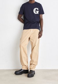 Camiseta de algodón navy con el logo blanco de "G" en el lado izquierdo del pecho, combinada con pantalones sueltos beige y zuecos de suela gruesa negros.