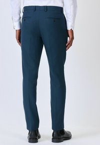 Tyrkysové kalhoty na míru vyrobené z texturované látky, disponující dvěma zadními kapsami s knoflíky, elegantně střiženými nohavicemi a nošené s černými botami.