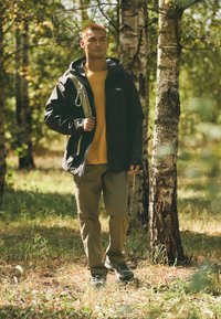 Czarna wodoodporna kurtka z regulowanym kapturem, żółta koszula pod spodem, zielone spodnie cargo i buty trekkingowe, usytuowane w lesie.