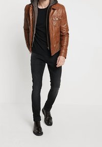 Bruine leren jas met ritssluiting over een zwart T-shirt, gecombineerd met zwarte jeans en glanzende zwarte enkellaarzen.
