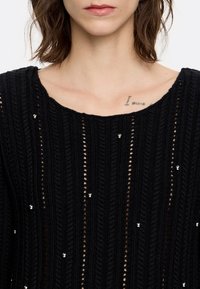 Schwarzer Strickpullover mit offener Maschenstruktur und vertikalem Rippenmuster, versehen mit kleinen, gleichmäßig verteilten silbernen Verzierungen.