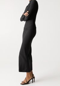 Robe noire cintrée à manches longues avec un détail froncé sur le côté, associée à des talons noirs à brides, mettant en valeur une texture lisse et une silhouette élégante.