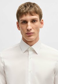 Camicia bianca in cotone con colletto rigido, chiusura con bottoni sul davanti e texture liscia. Nessun motivo o logo visibile.