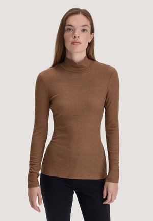 Jeune femme aux cheveux lisses châtain clair porte un col roulé côtelé marron ajusté et un pantalon noir, debout devant un fond uni.