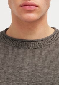 Pull en maille tricotée gris foncé avec un col rond côtelé et une texture lisse, présentant une vue rapprochée du col et de la partie inférieure du visage.