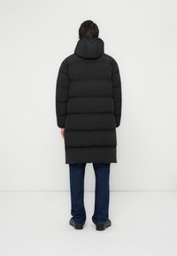 Zwarte pufferjas met capuchon, voorzien van een gestikt ontwerp en een verlengde vorm. Gepaard met blauwe jeans en zwarte schoenen.