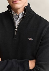Hombre con suéter negro con cremallera hasta el cuarto, sobre una camisa de cuadros con botones, con los brazos cruzados, mostrando un pequeño logo bordado en el pecho.
