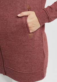 Burgunderfarget hettegenser med glidelås, med myk stofftekstur, skrå lommer med brune trekantede detaljer, og ribbede mansjetter på ermene.