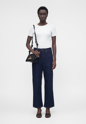 Femeie care poartă un top alb cu mâneci scurte și blugi evazați în culoare închisă, ținând o geantă neagră de umăr cu logo auriu, stând în fața unui fundal simplu.