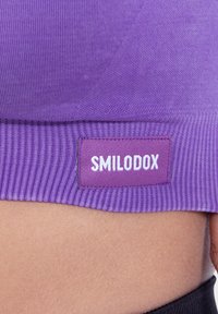Violetti ribattu kangas urheilu paita, jossa on valkoinen SMILODOX-tunnus violetilla taustalla helman kohdalla. Paidassa on sileä pinta.
