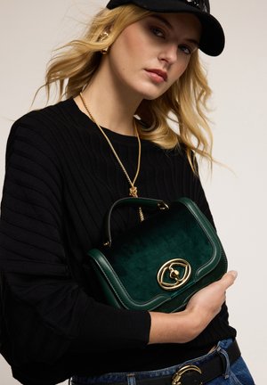 Borsa in velluto verde con una chiusura ovale dorata, caratterizzata da una forma strutturata e un manico corto, tenuta da una persona che indossa un maglione nero a coste.