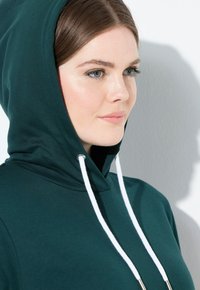 Mujer joven con piel clara y ojos azules, vistiendo una sudadera verde oscura con cordones blancos, mirando hacia la derecha contra un fondo blanco.
