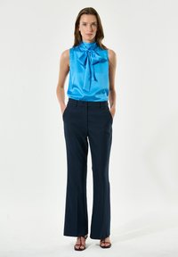 Blusa azul de satén con cuello alto y detalle de lazo, combinada con pantalones de sastre en navy. El conjunto presenta una silueta elegante y fluida.