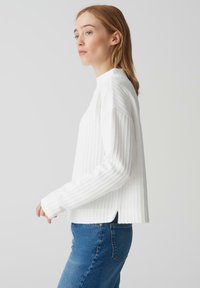 Weißer gerippter Pullover mit hohem Kragen, langen Ärmeln und seitlichen Schlitzen. Kombiniert mit blauen Jeans, die eine lässige Silhouette betonen.