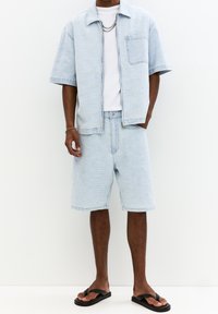 Světle modrá denimová košile s krátkými rukávy a kapsou, doplněná o sladěné šortky. Doplňky zahrnují stříbrný řetízek a černé žabky.