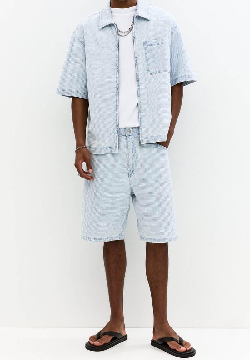 Světle modrá denimová košile s krátkými rukávy a kapsou, doplněná o sladěné šortky. Doplňky zahrnují stříbrný řetízek a černé žabky.