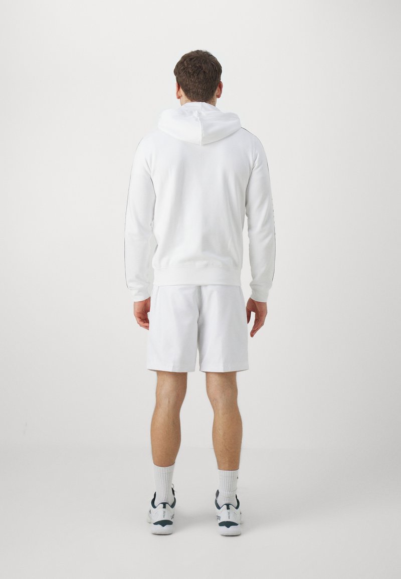 Felpa bianca con orlo e polsini a costine, abbinata a pantaloni corti bianchi. Dettagli di cucitura evidenti sulle maniche e una texture del tessuto confortevole.