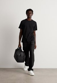 Man in zwart t-shirt en broek draagt een zwarte sporttas in zijn rechterhand, terwijl hij over een grijze vloer loopt tegen een eenvoudige witte muur.