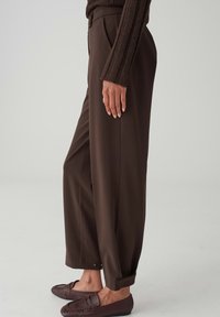 Vrouw die donkerbruine broek met wijde pijpen, bijpassende loafers en een geribbelde trui met lange mouwen draagt, zijwaarts staand tegen een effen achtergrond.