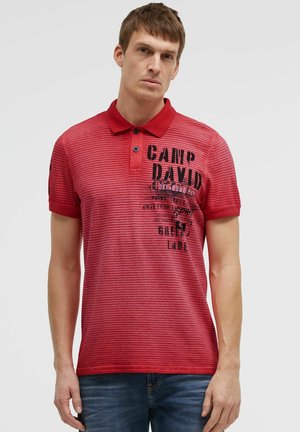 Man wearing red striped polo shirt with black text on the chest and blue jeans, standing against plain light background.