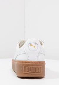 Zapatilla blanca de cuero con suela de goma, que presenta un logo dorado, detalles cosidos y un cuello acolchado para mayor comodidad.
