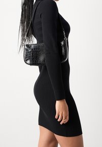 Borsa nera con motivo coccodrillo e tracolla, abbinata a un vestito aderente nero a costine, che mette in mostra una silhouette slanciata.
