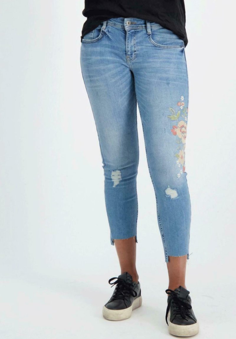 Blauwe denim 3/4 jeans met bloemborduurwerk op de rechter bovenbeen, versleten details, onafgewerkte zoom en een slim fit. Draagt met zwarte sneakers.