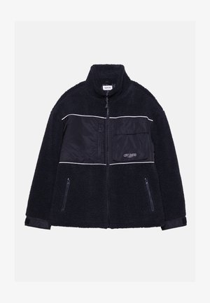 Μαύρο φλις μπουφάν με ψηλό γιακά, αντίθετο λείο πάνελ υφάσματος, τσέπες με φερμουάρ και λευκές λεπτομέρειες. Υφή ζεστή και άνετη.