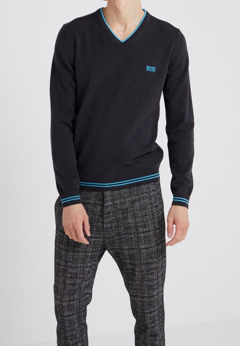 Homme portant un pull noir à col en V avec une bordure bleue et un logo, associé à un pantalon gris à motifs sur un fond clair uni.