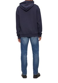 Hoodie van donkerblauwe stof, relaxed fit, kangoeroezak zichtbaar aan de voorkant, gedragen met lichtblauwe jeans en zwarte schoenen.
