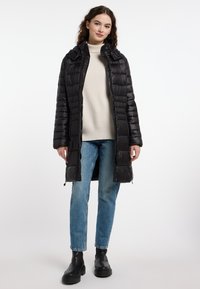 Zwarte lange pufferjas met ritssluiting, voorzien van een capuchon en geribde manchetten, gedragen over een lichte trui en blauwe jeans. Zwarte laarzen, eenvoudig ontwerp.