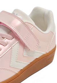 Lyserøde sneakers med en blank finish, der har et hvidt zigzag-design, en velcrolukning og en brun gummisål. Glat tekstur og afrundet form.