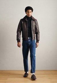 Hnedá kožená bomber bunda s kožušinovým golierom, kombinovaná s tmavomodrými džínsami a hnedými topánkami, nosená cez čierny rolák.