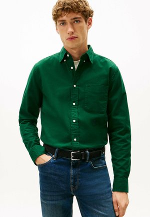 Camisa verde de botones hecha de algodón, con un bolsillo en el pecho y puños abotonados, combinada con unos vaqueros de mezclilla azul y un cinturón negro trenzado.