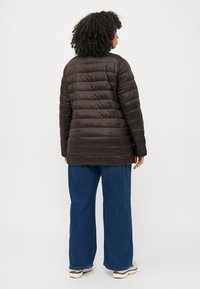 Brown puffer jacket with horizontal quilting, featuring a high collar, soft texture, and a slightly longer cut. Paired with wide denim pants.