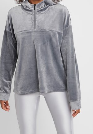 Vrouw draagt een zachte grijze hoodie met een rits aan de voorkant en een losse pasvorm, gecombineerd met glanzende witte legging tegen een effen achtergrond.