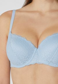 Sujetador de encaje de color azul claro con bordes festoneados, soporte de aros y tirantes ajustables. Presenta patrones florales y una textura suave.