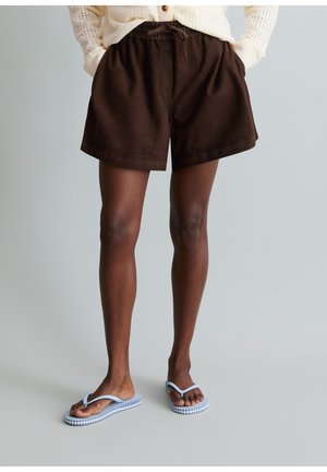 Persoon draagt bruine short met trekkoord, witte geperforeerde trui en lichtblauwe teenslippers, staand op een lichtgrijze vloer.
