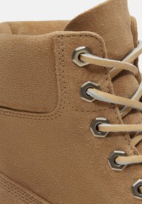 Primo piano di stivale in camoscio color nocciola con spesse stringhe beige infilate attraverso occhielli esagonali in metallo su colletto imbottito alla caviglia.