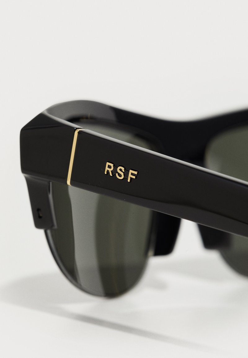 Zwarte zonnebril met groene lenzen, voorzien van een glanzende afwerking, gouden accenten en "RSF" branding op de tempels. Geometrisch frameontwerp.
