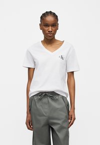 Γυναίκα με πλεξούδες, που φοράει λευκό T-shirt Calvin Klein με V-λαιμόκοψη και γκρι παντελόνι με κορδόνι, στέκεται μπροστά σε απλό φόντο.