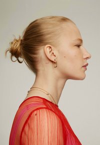 Vista de perfil de una persona con un peinado de moño. Lleva una blusa roja transparente, con cadenas de oro en capas y pendientes de aro. Tatuaje notable en el cuello.