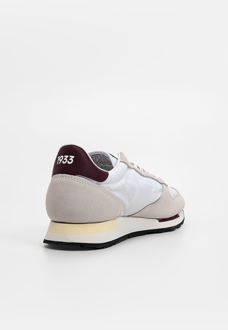 Zapatilla con una parte superior de nailon blanco, acentos de ante beige, talón color marrón y suela color crema. Presenta un prominente "1933" en el talón.