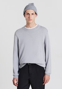 Maglione a maglia grigio chiaro con collo rotondo e polsini a coste, abbinato a pantaloni scuri. Un berretto grigio abbinato completa il look.