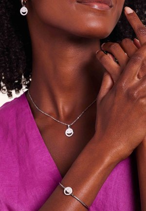 Ensemble de bijoux en argent comprenant un pendentif en forme de visage souriant circulaire sur une délicate chaîne de collier et un bracelet assorti ; le modèle porte un haut violet vif.