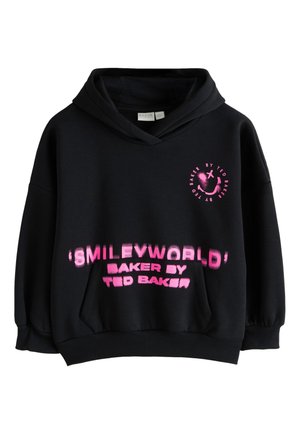 Ragazza adolescente che indossa una felpa nera oversize con faccina rosa sorridente e la scritta "Baker by Ted Baker", in piedi contro una parete chiara e semplice.